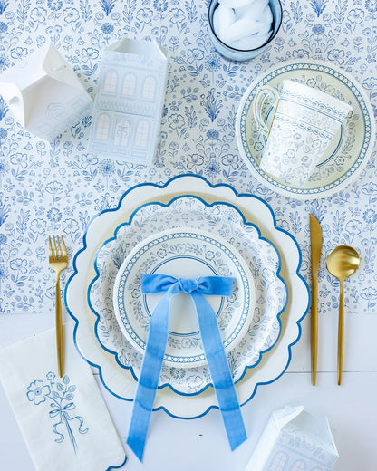 Table set with blue and white patterned paper plates, matching paper teacup, gold cutlery, and floral napkin. Background features a floral table runner, with a blue ribbon bow atop the plate setting.