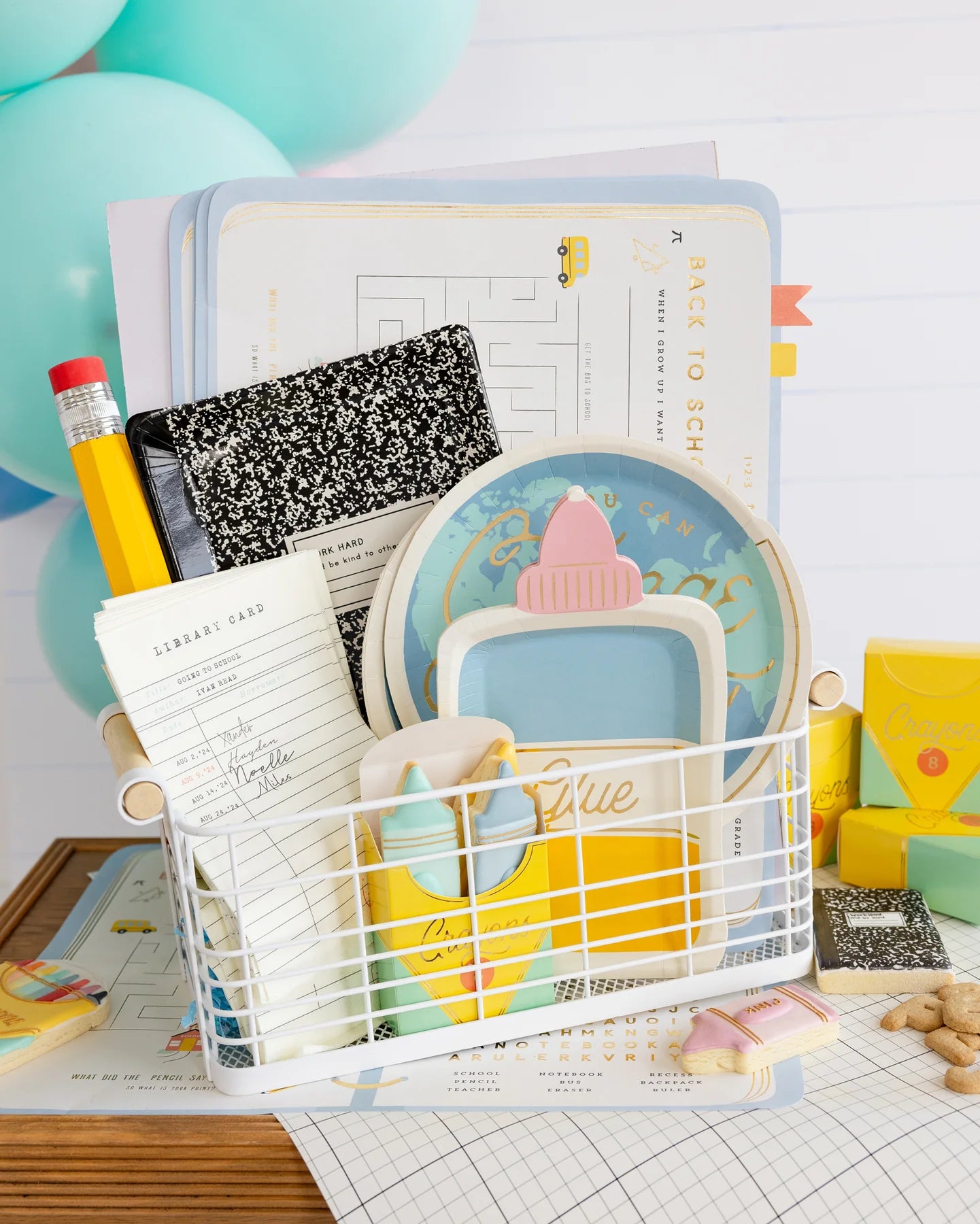 A wire basket holding back to school themed party supplies including notebook paper plate, globe shaped paper plate and crayon treat boxes