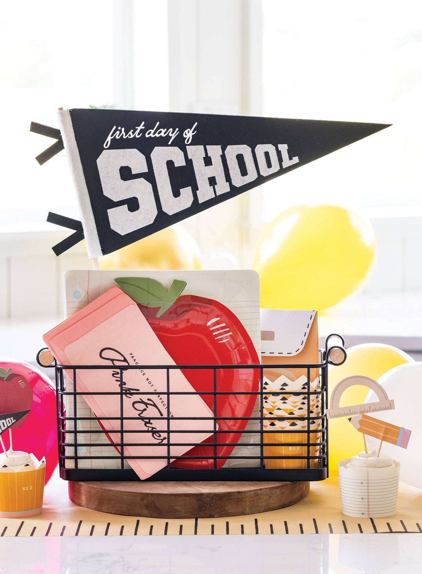 a basket filled with first day of school party supplies including a pennant flag, apple shaped paper plate and notebook inspired paper plates