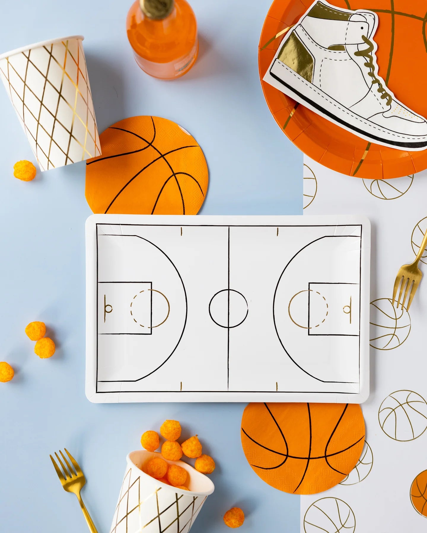 A party setup with a basketball theme includes a rectangular paper plate featuring a basketball court design, orange basketball-shaped plates and napkins, a cup of cheese puffs, a bottle of orange soda, a sneaker illustration on a napkin, and gold plastic utensils.