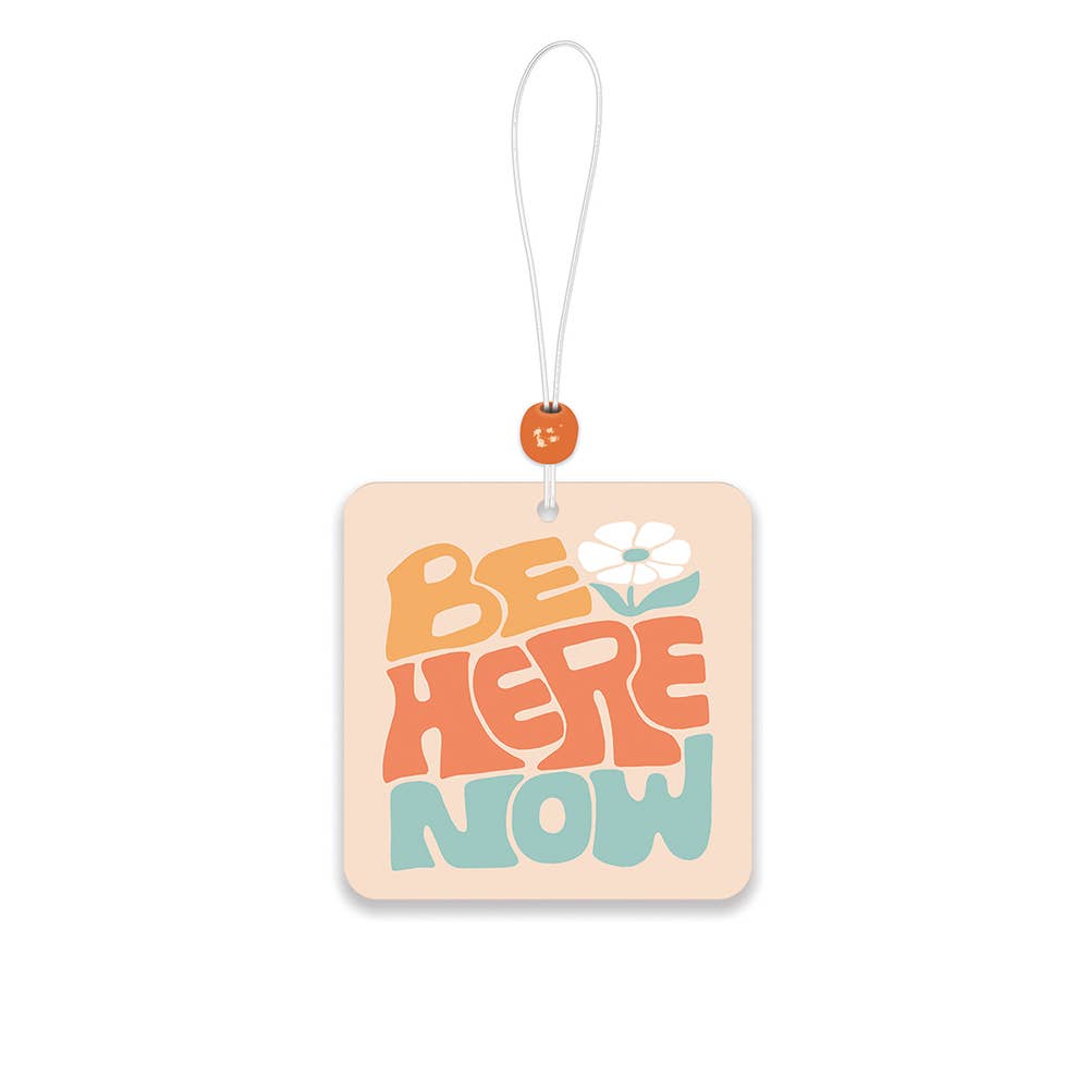 square "Be Here Now" car air freshener with a flower design 