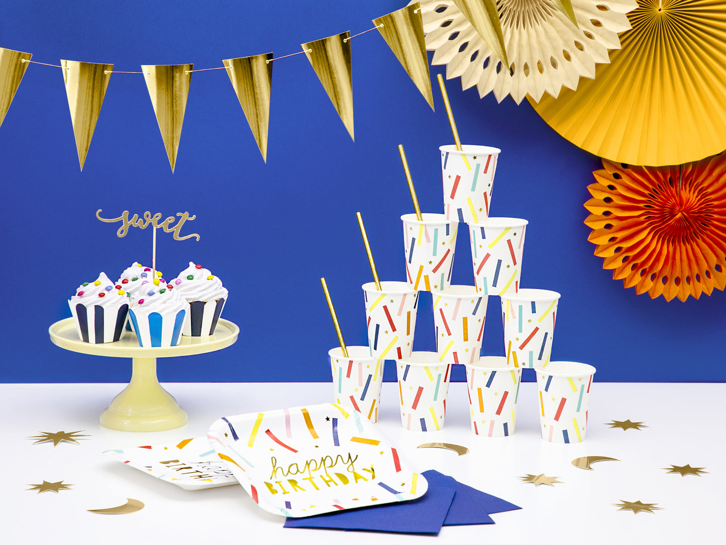 Happy birthday table with a tower of colourful plates and cups, a cake stand with cupcakes and toppers, and a hanging pennant banner and party fans in the background.