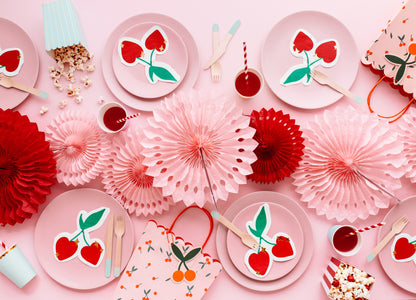 Flat lay of cherry-themed party setup with pink paper fans, cherry napkins, and assorted decorations.