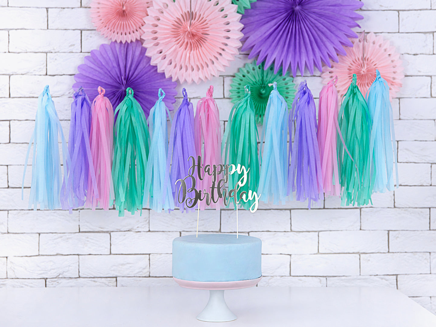 Birthday cake with a silver "Happy Birthday" topper in front of a backdrop of purple, pink, green, and blue streamers and paper fans.