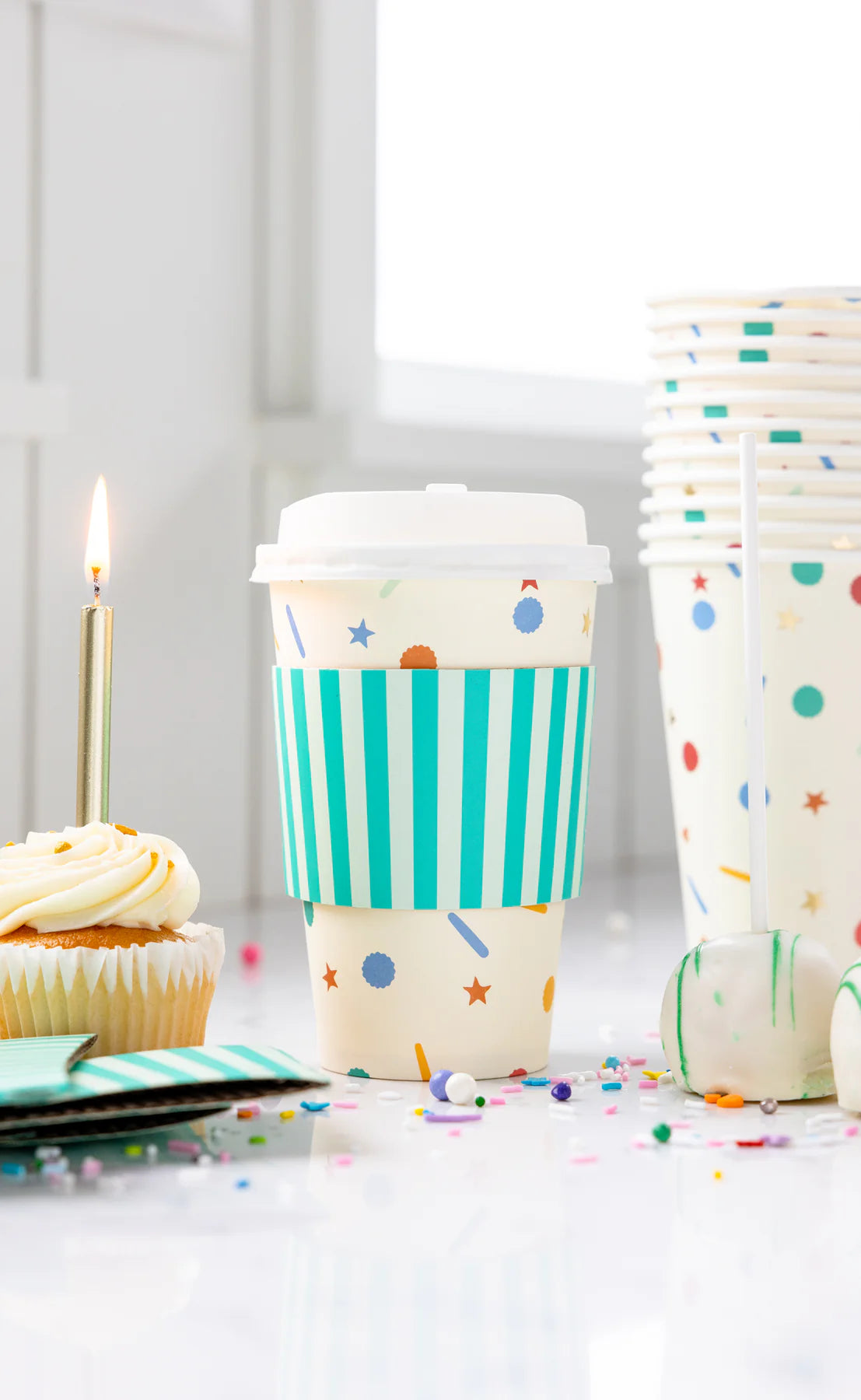 Confetti-designed paper to-go cup with plastic lid and teal striped sleeve on white counter, next to stacked cups and cupcake with lit candle, surrounded by confetti.