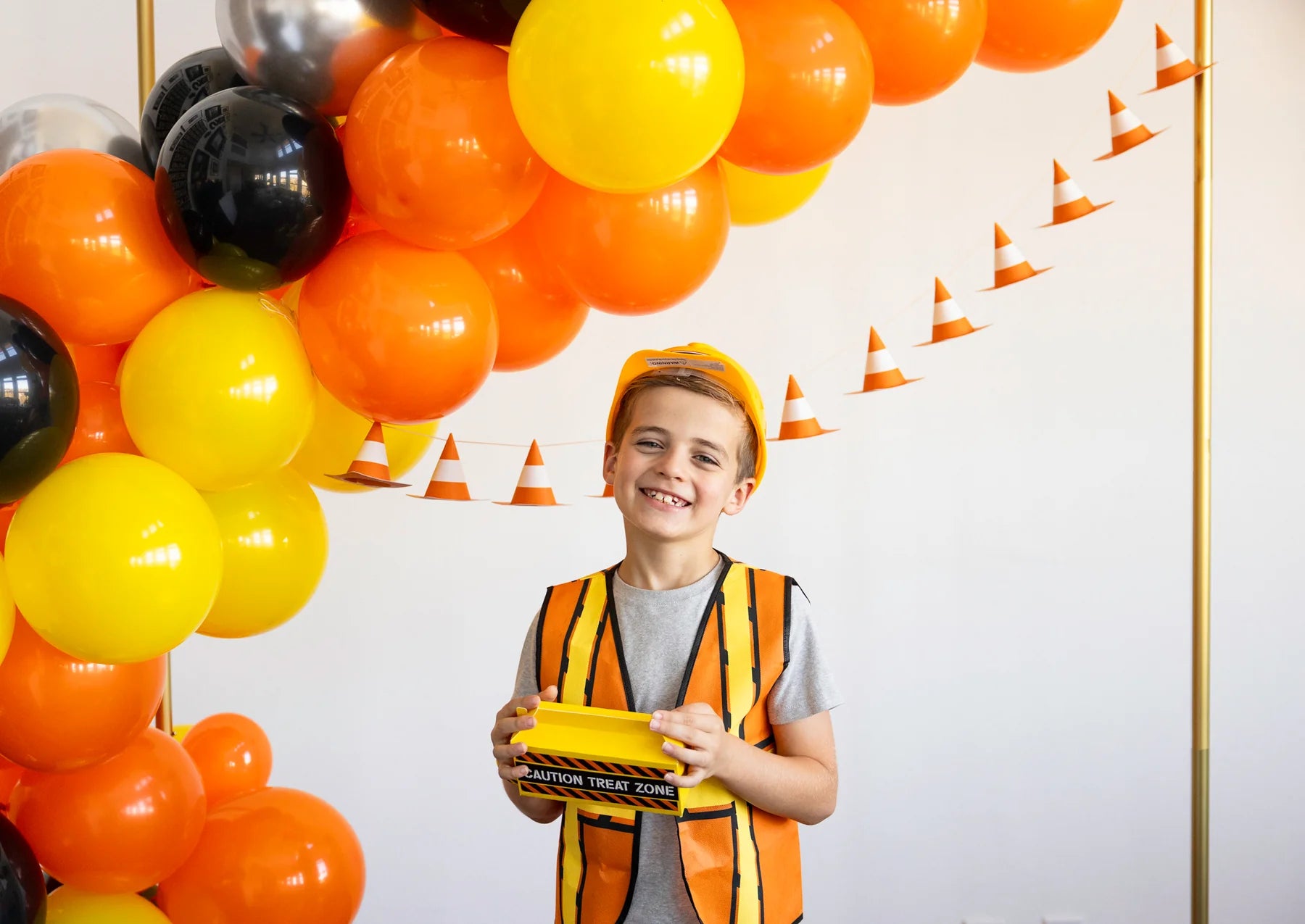 Young boy in a construction costume posing under an orange and yellow balloon garland.