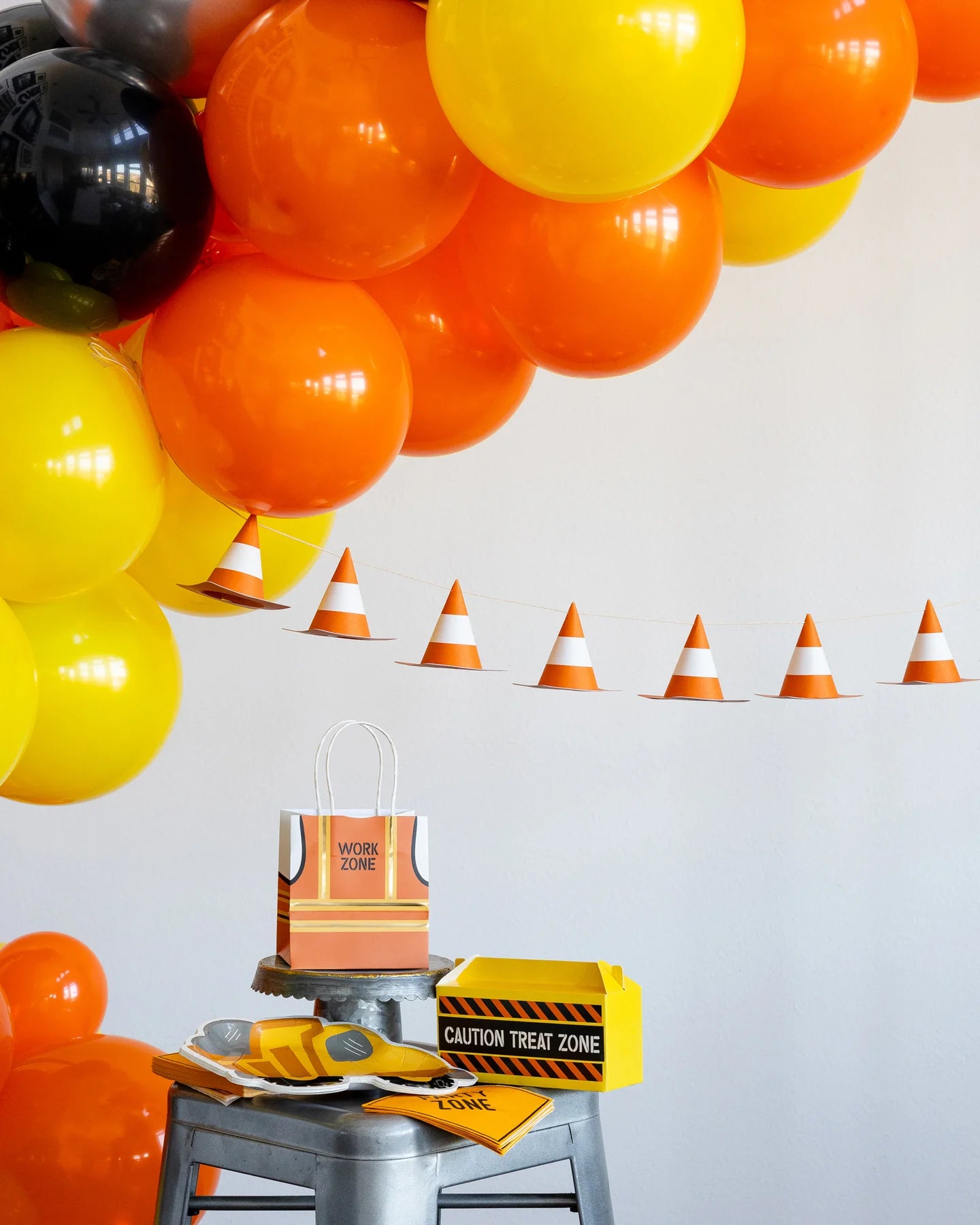 Orange and yellow balloons with a mini traffic cone banner and construction themed party decorations on a gray stool.