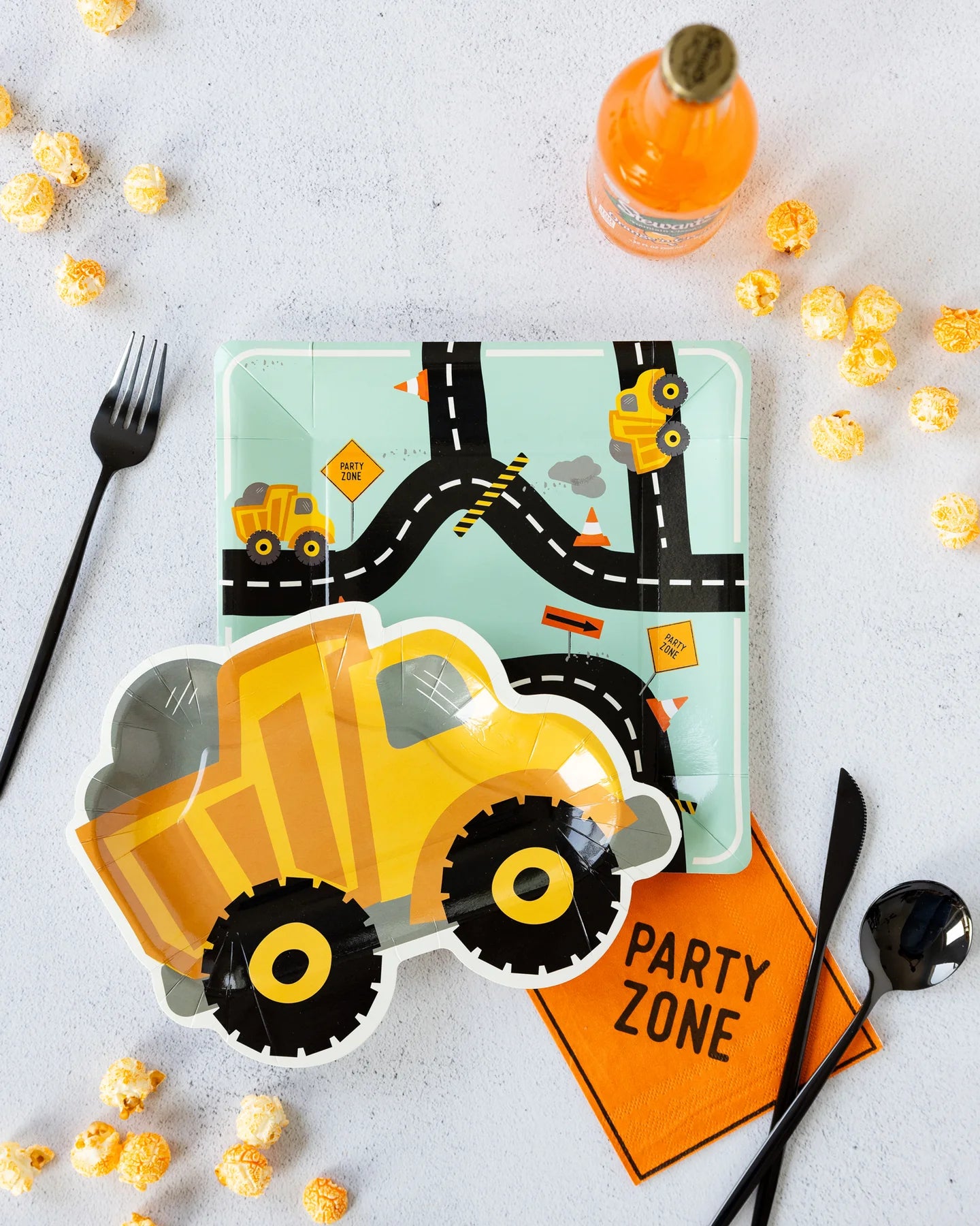 Party table setting with construction-themed plates, fork, spoon, popcorn, orange napkin, and orange soda bottle.