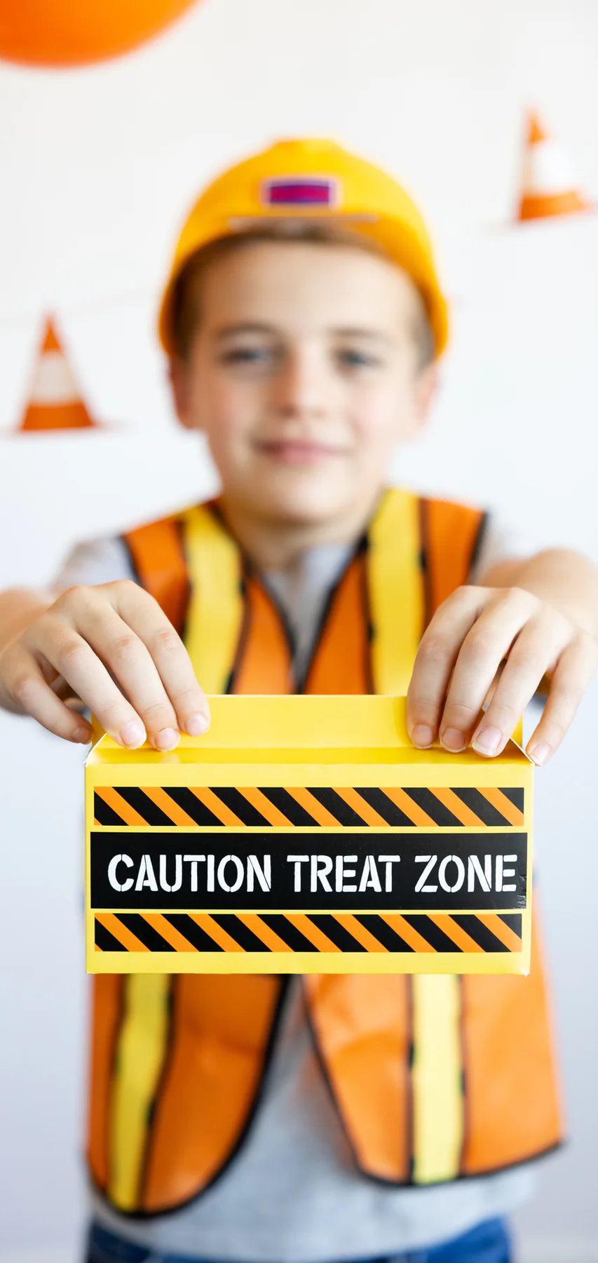 Child in a construction outfit holding a box labeled "Caution Treat Zone," with traffic cone decor in the background.