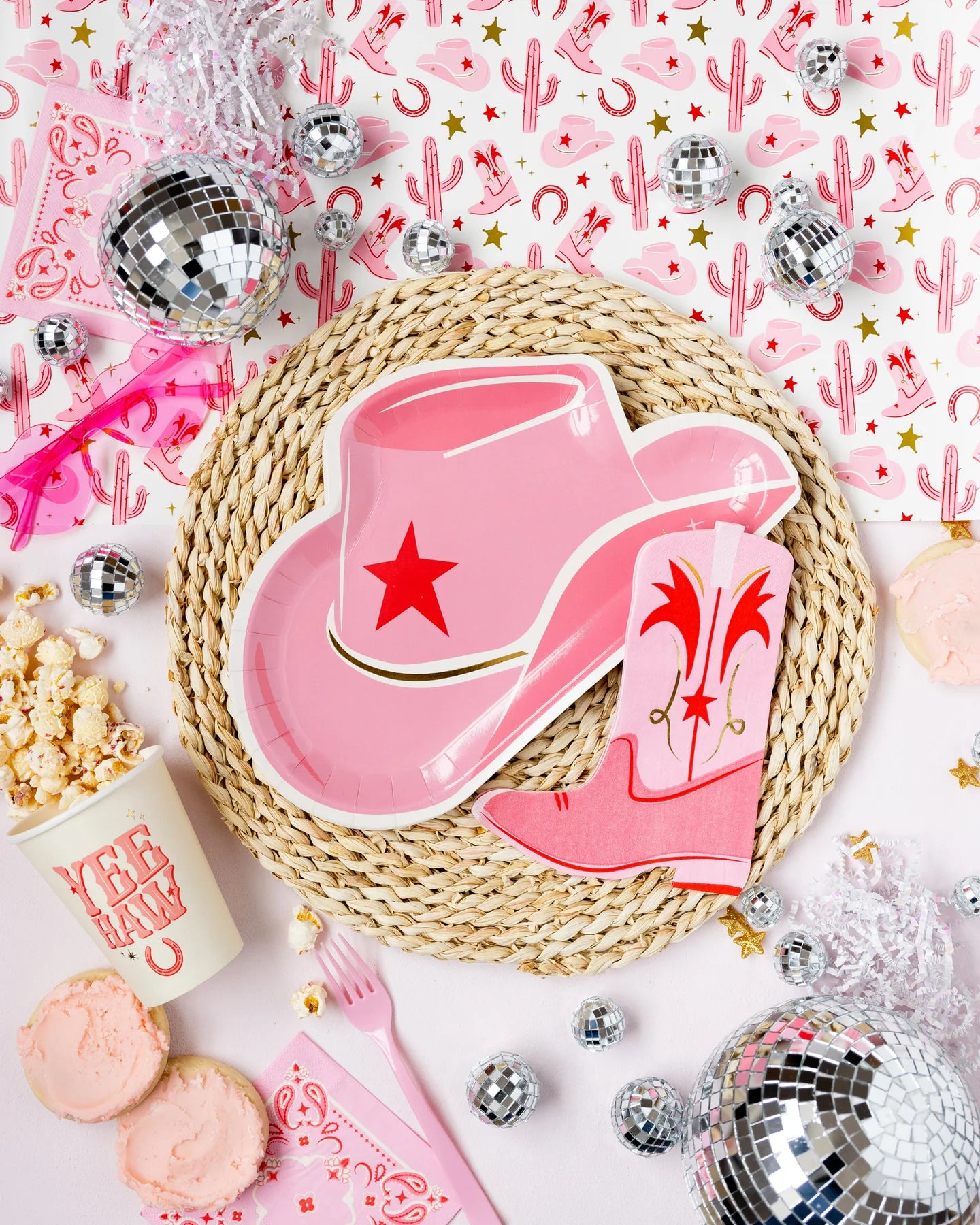 Western-themed party decor with a pink cowboy hat paper plate, boot napkin, disco balls, and popcorn on a woven mat.