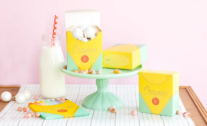 Crayon treat boxes on a pastel green cake stand with a bottle of milk and a pink straw, against a pink background.