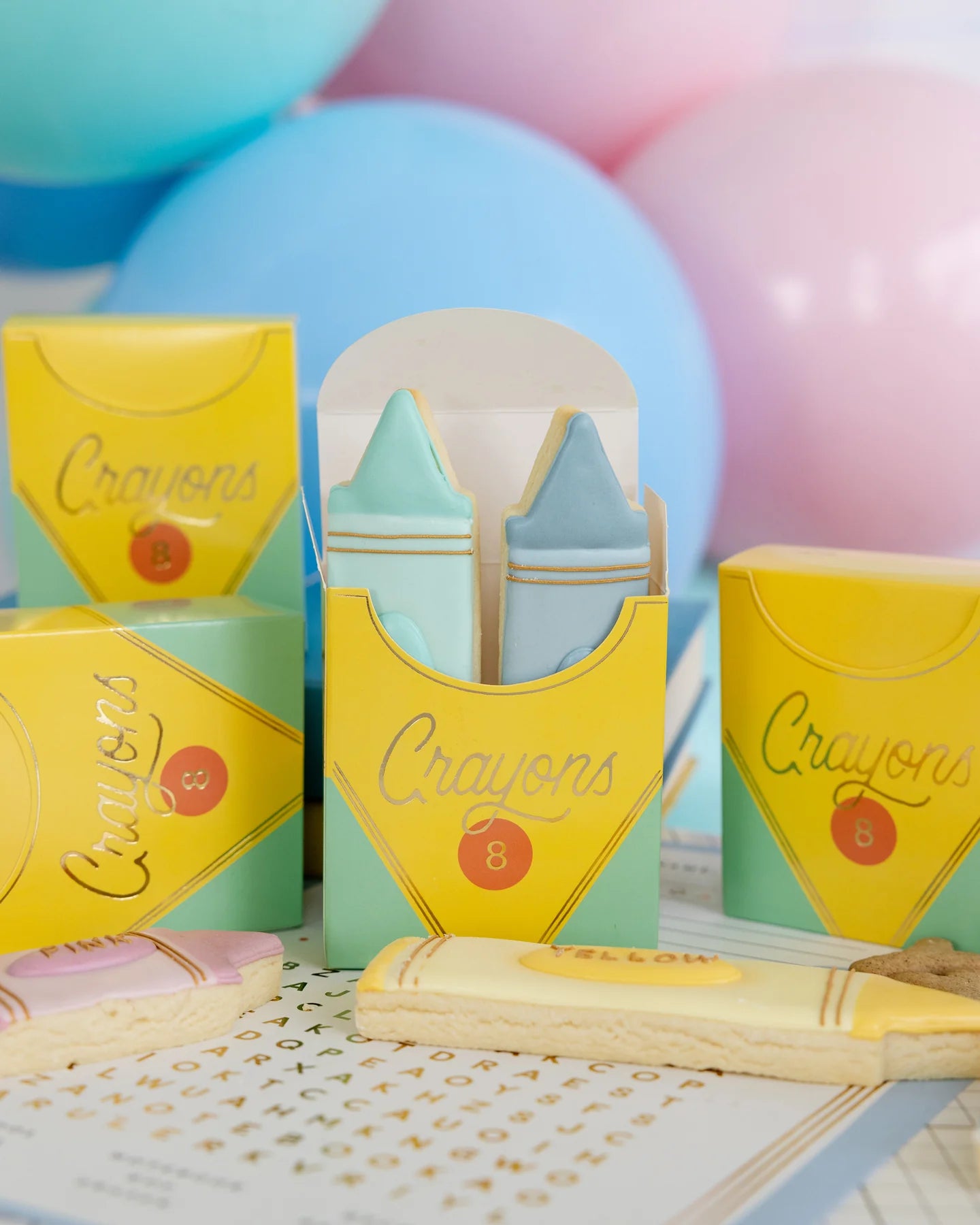 Colorful crayon-shaped cookies in yellow crayon-themed boxes with pastel balloons in the background.