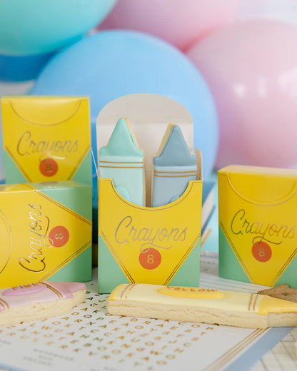 Colorful crayon-shaped cookies in yellow crayon-themed boxes with pastel balloons in the background.