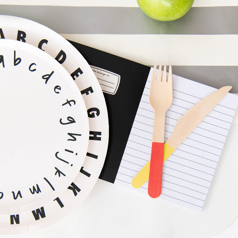 Alphabet-themed dessert and dinner plates on a school notebook, with a wooden fork and knife with coloured ends beside them