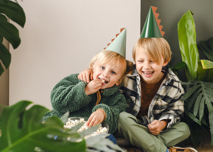 two friends sitting down eating popcorn and wearing the dinosaur party hats