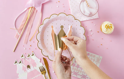 Hands decorating a pink and gold crown on a plate surrounded by princess-themed party supplies including a cupcake, a paper castle, a pink star wand, and gold utensils on a pink tablecloth.