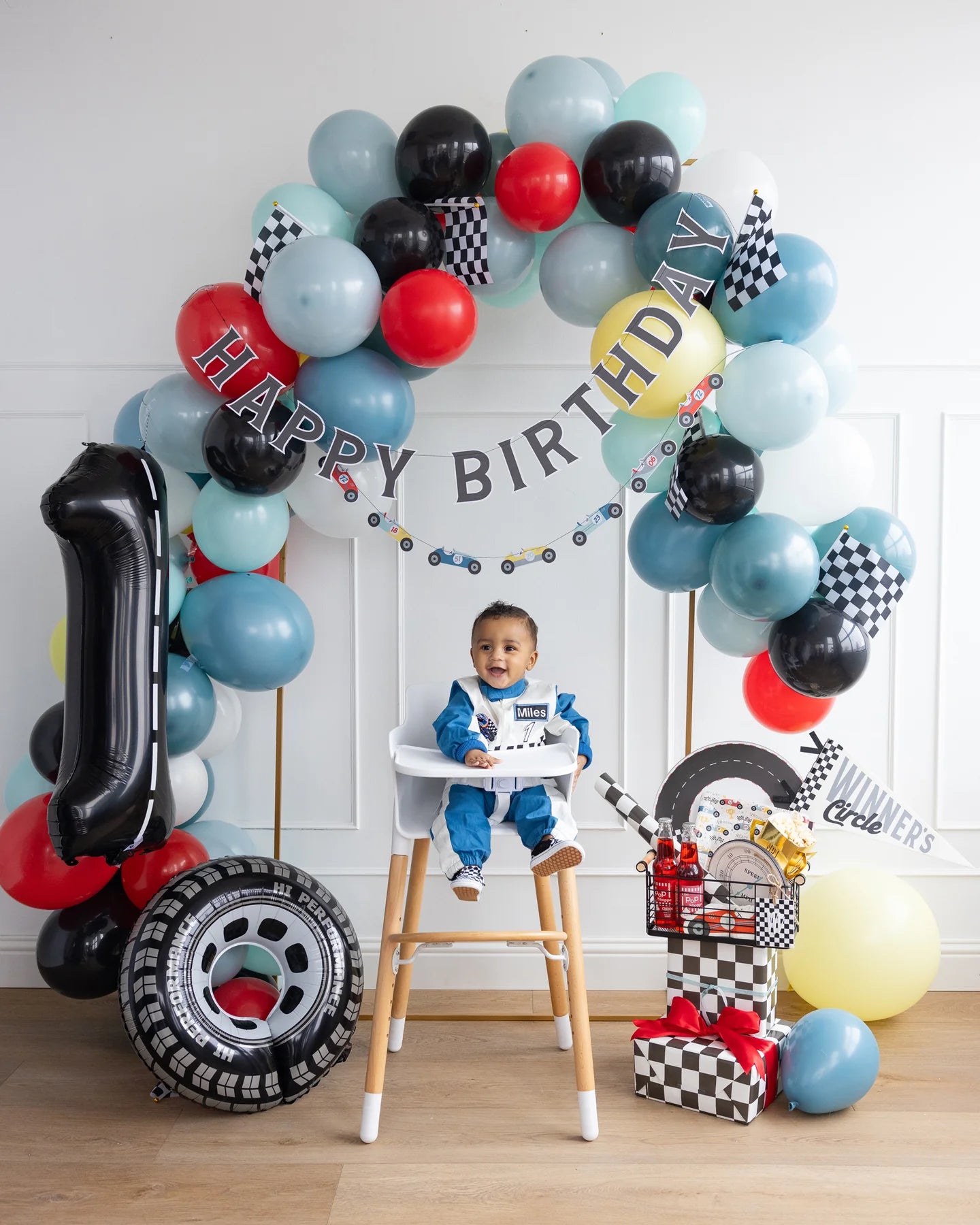 Baby in a high chair at a racing-themed birthday party with balloons, checkered flags, and gift-wrapped presents.