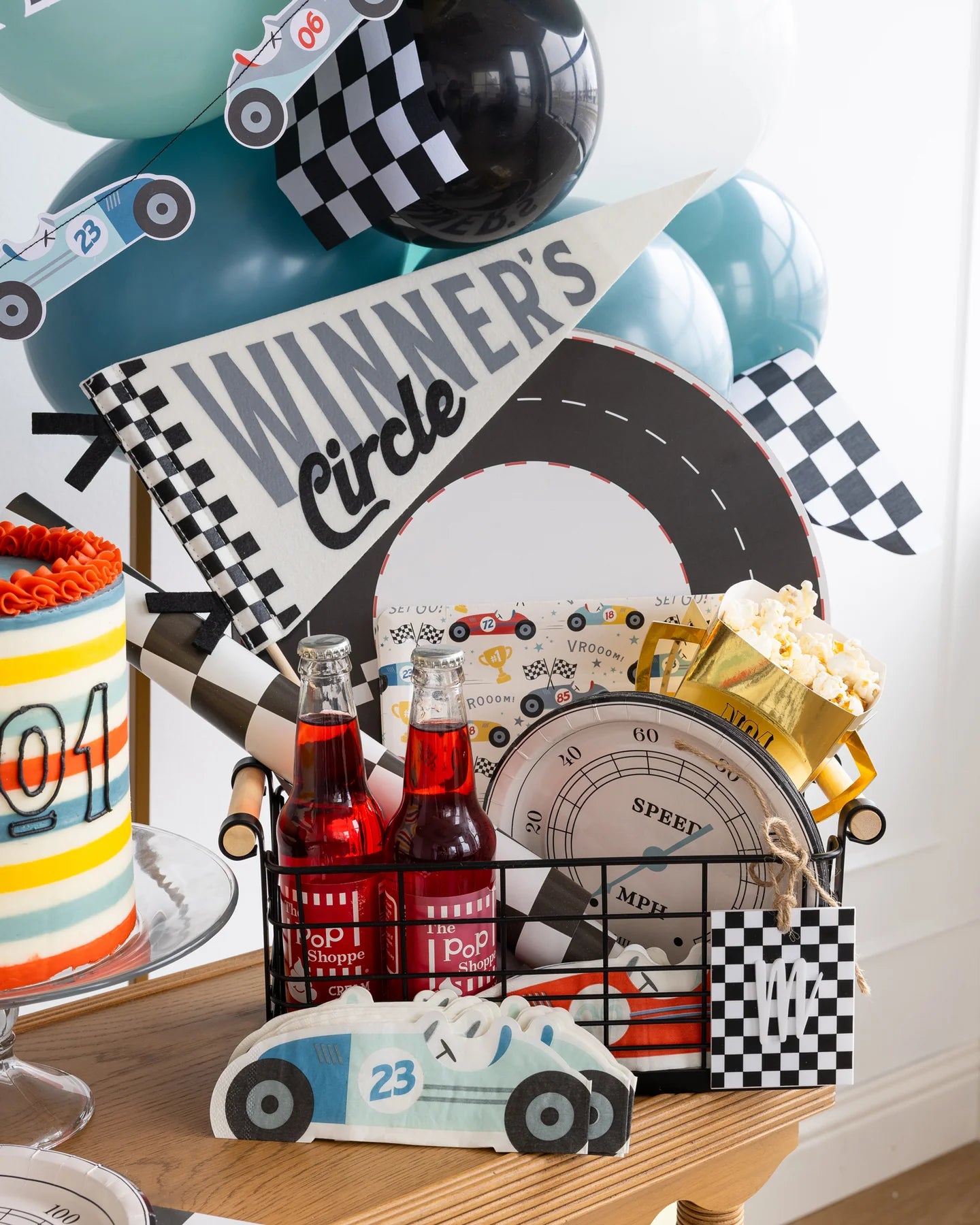 black wire  basket filled with race car themed party supplies including a speedometer paper plate, 'winners circle' pennant flag and red soda bottles 