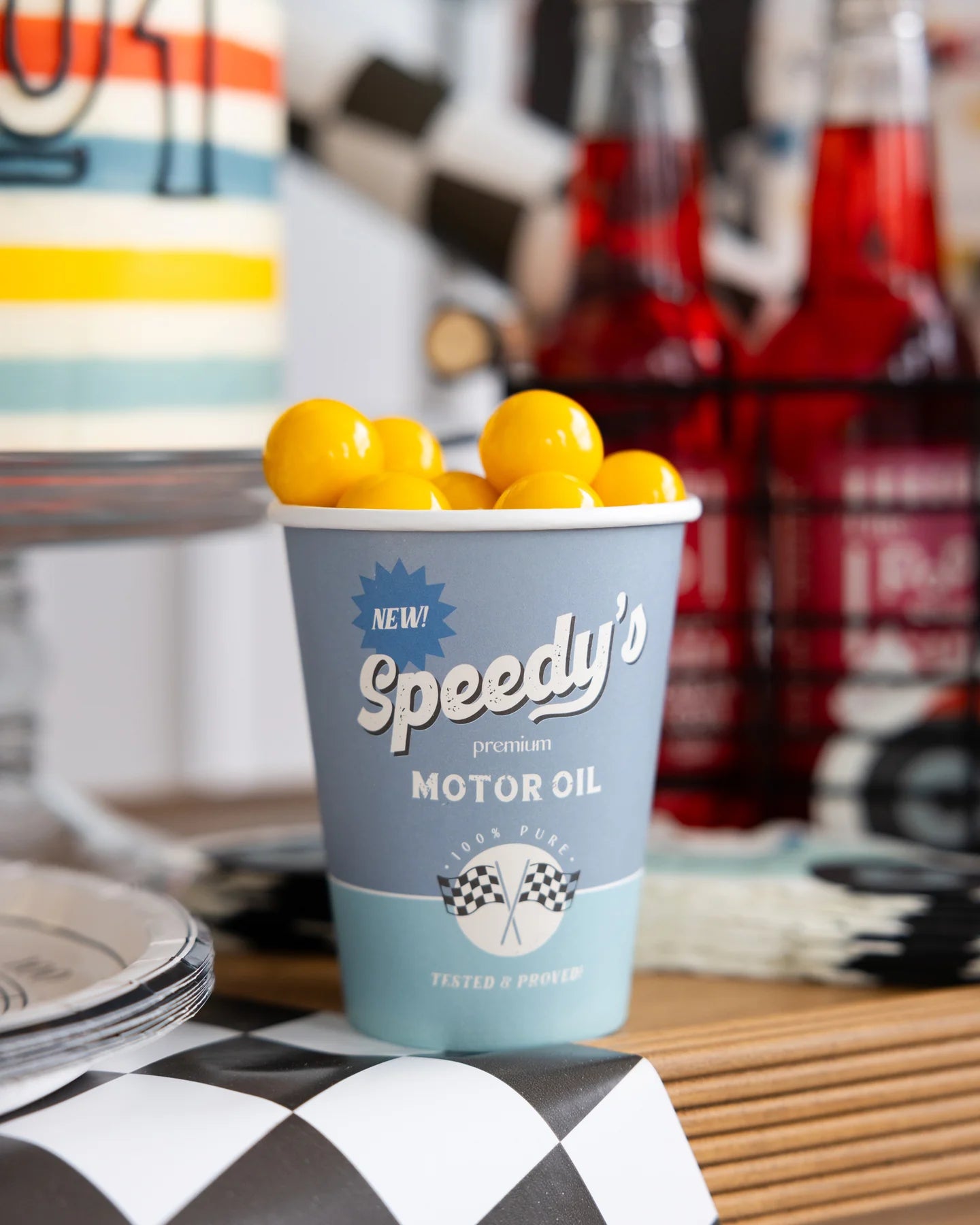 Light blue disposable cup filled with yellow gumball candies. Features 'Speedy's Premium MOTOR OIL' in white text, '100% PURE! TESTED & PROVED!' with crossed checkered flags, and a 'NEW!' blue burst.