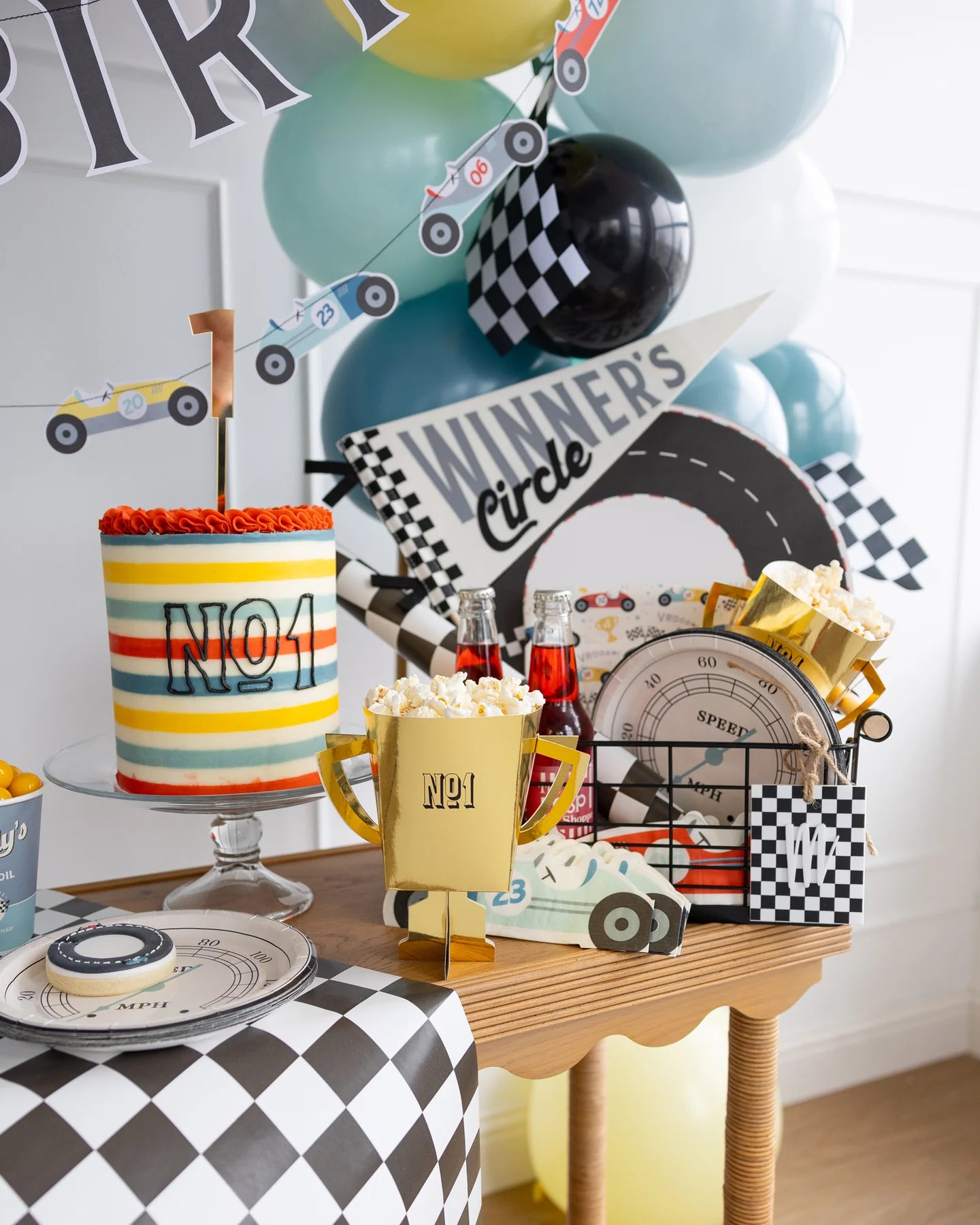 Race car-themed decorated table with striped cake topped with number 1, 'Winner's Circle' sign, trophy treat boxes with popcorn, soda bottles, balloons, checkered and car-themed decorations.