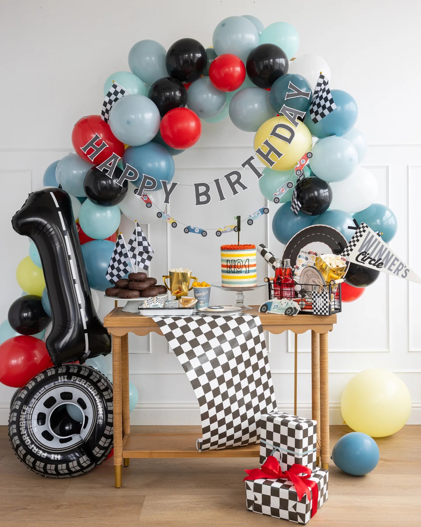 1st birthday dessert table setup with race car inspired party supplies, a balloon garland and wrapped presents.