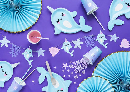 flat lay photo of narwhal themed paper party supplies including banner, plates, cups and party fans