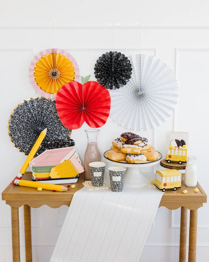 Back-to-school themed table setup with colourful paper fans on wall, notebooks, pencils, paper cups, chocolate milk jug, milk bottle, pastries, and paper school bus decorations.