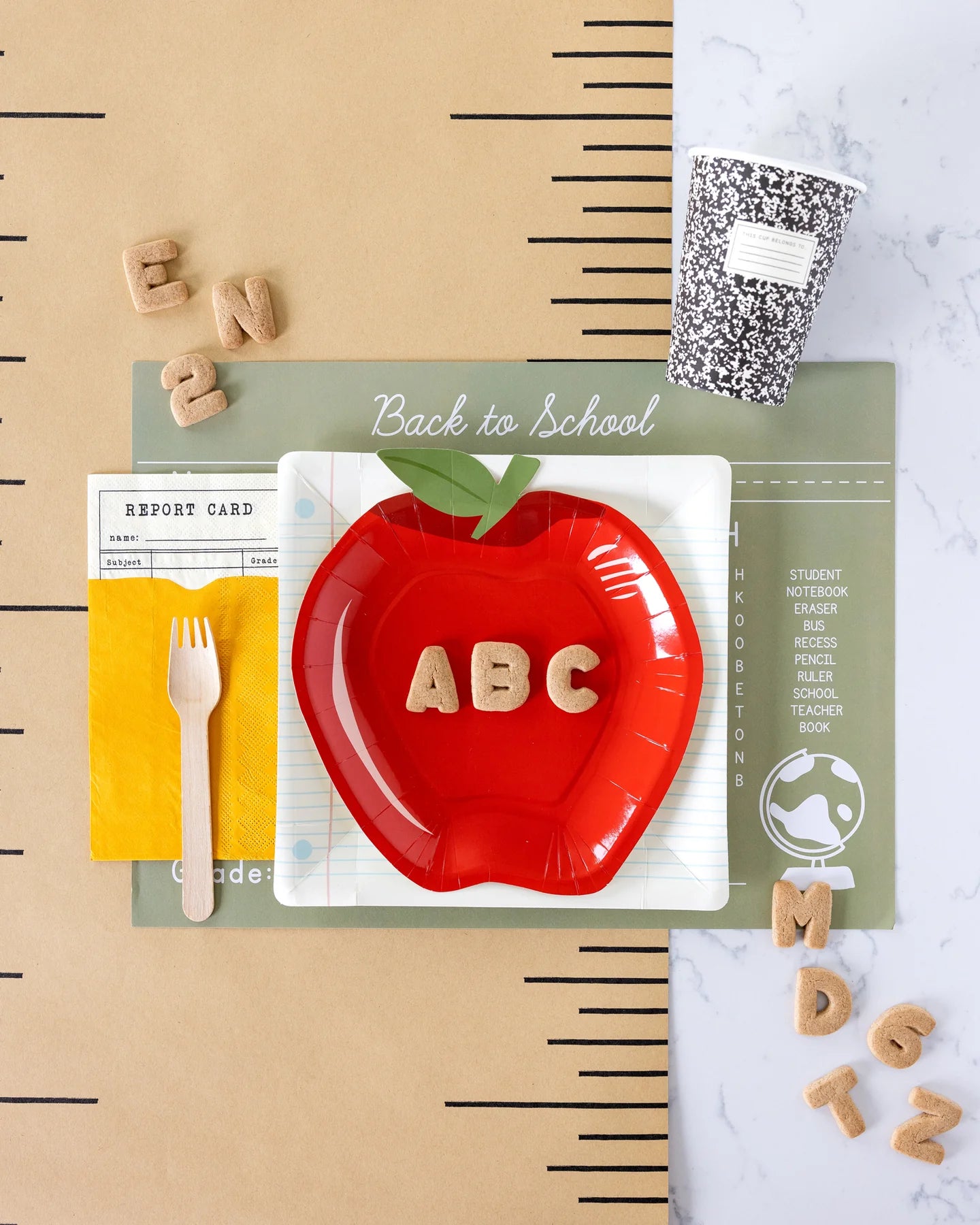 a flat lay photo with a back to school themed table setting including report card inspired napkins, apple shaped paper plate and composition styled paper cup