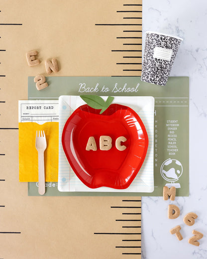 a flat lay photo with a back to school themed table setting including report card inspired napkins, apple shaped paper plate and composition styled paper cup