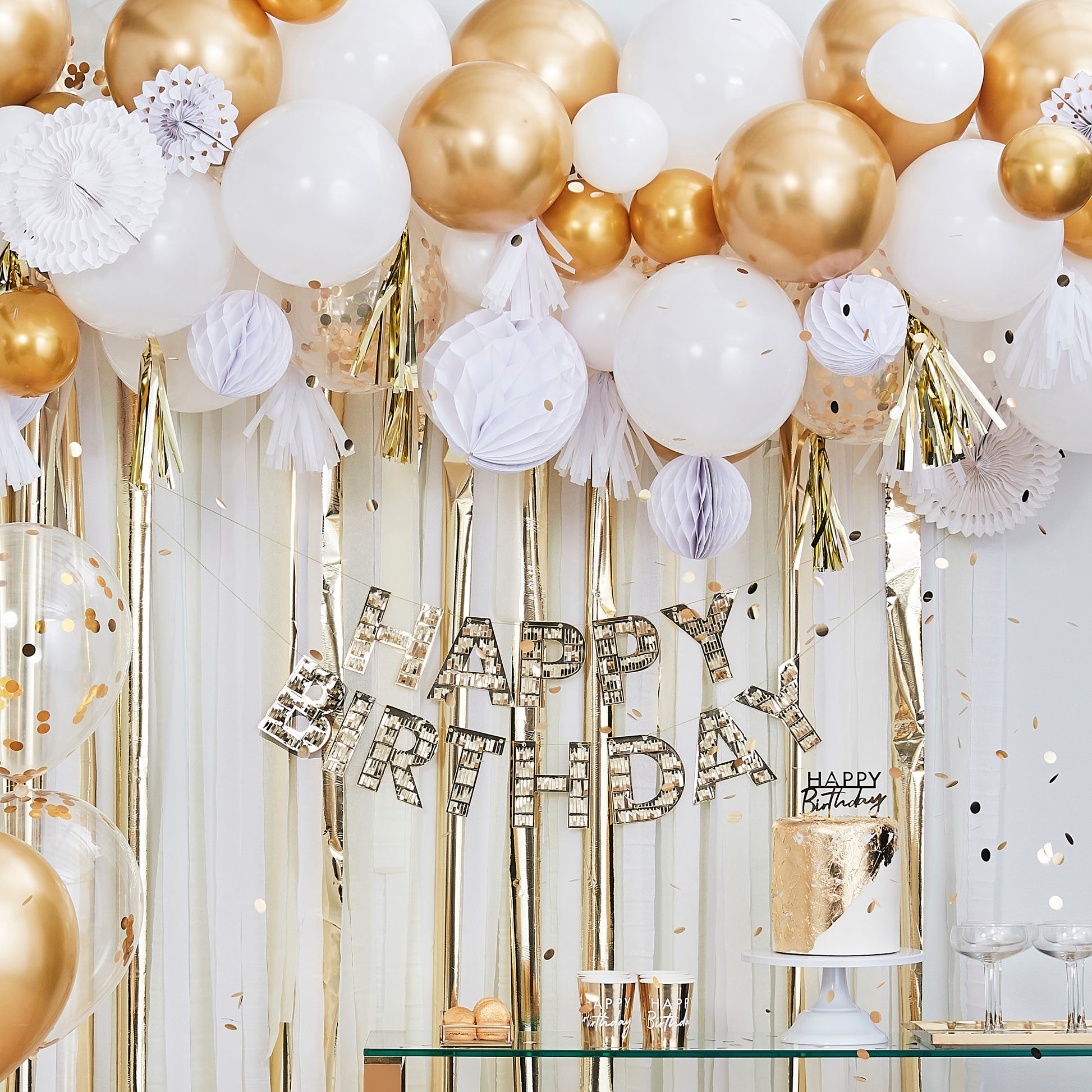 Birthday party setup with white and gold decor, streamer backdrop, hanging sign, and balloons.