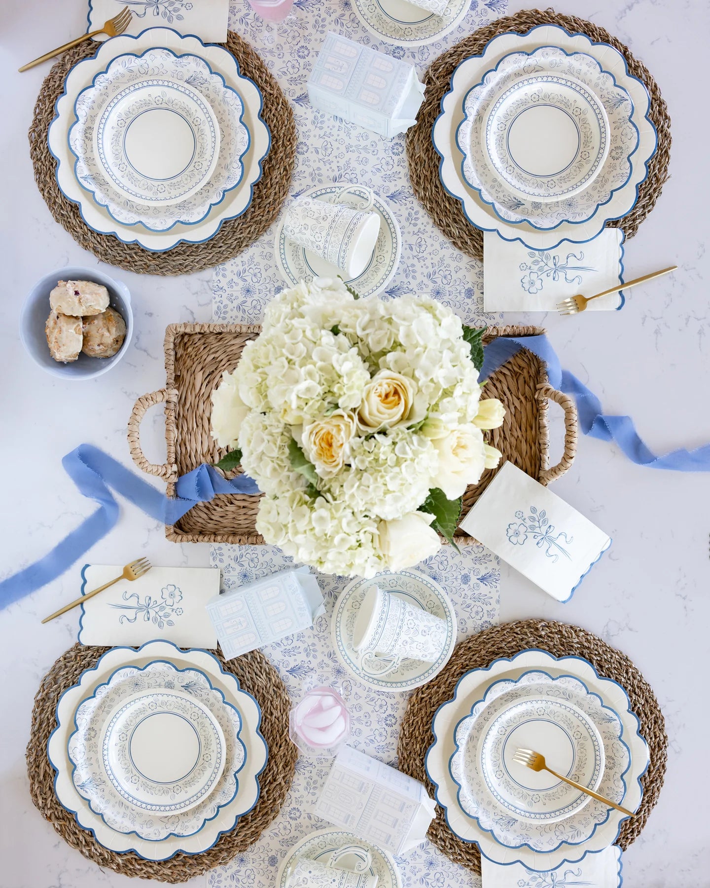 Table for four with blue and white floral napkins, paper plates, and cups. Centerpiece of white and cream flowers on a straw tray, gold cutlery, and small pastries in a bowl.