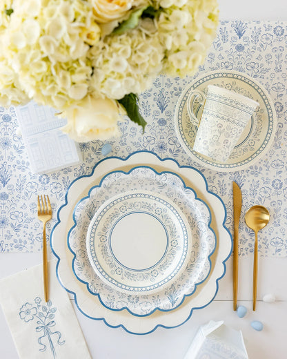 Table set with blue and white patterned paper plates, matching paper teacup, gold cutlery, and floral napkin. Floral table runner in background with bouquet of white flowers at top left.