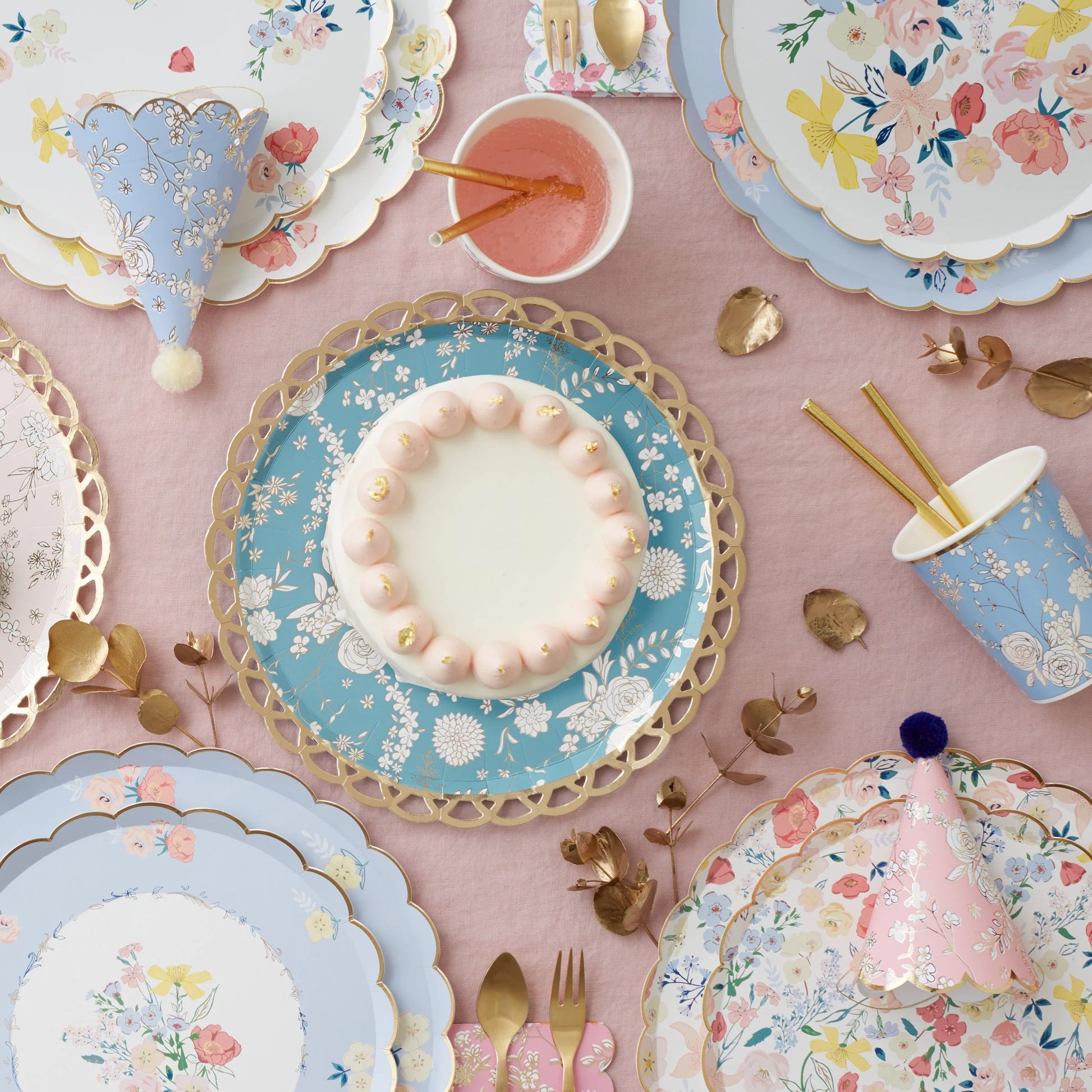 A table topped with floral themed plates, cups and party hats with a cake in the center.