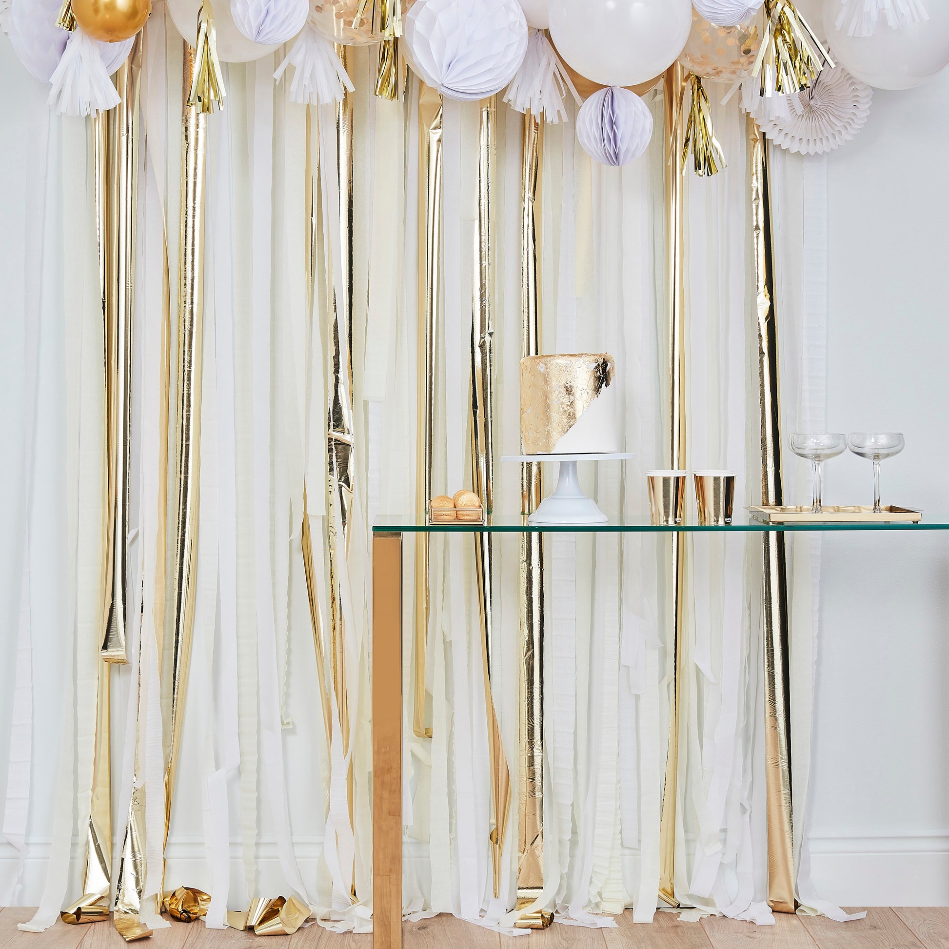 Birthday party setup with a table featuring cake and cups, set against a white and gold streamer backdrop.