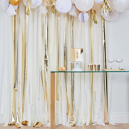 Birthday party setup with a table featuring cake and cups, set against a white and gold streamer backdrop.